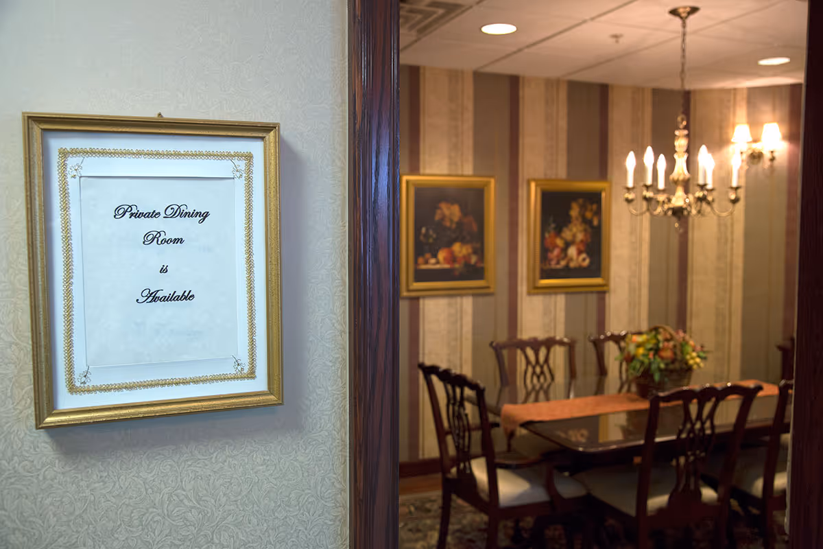 View into a private dining room with a wooden dining table, six chairs, a chandelier, and two framed paintings on the wall. A framed sign on the wall outside the room reads 'Private Dining Room is Available'.