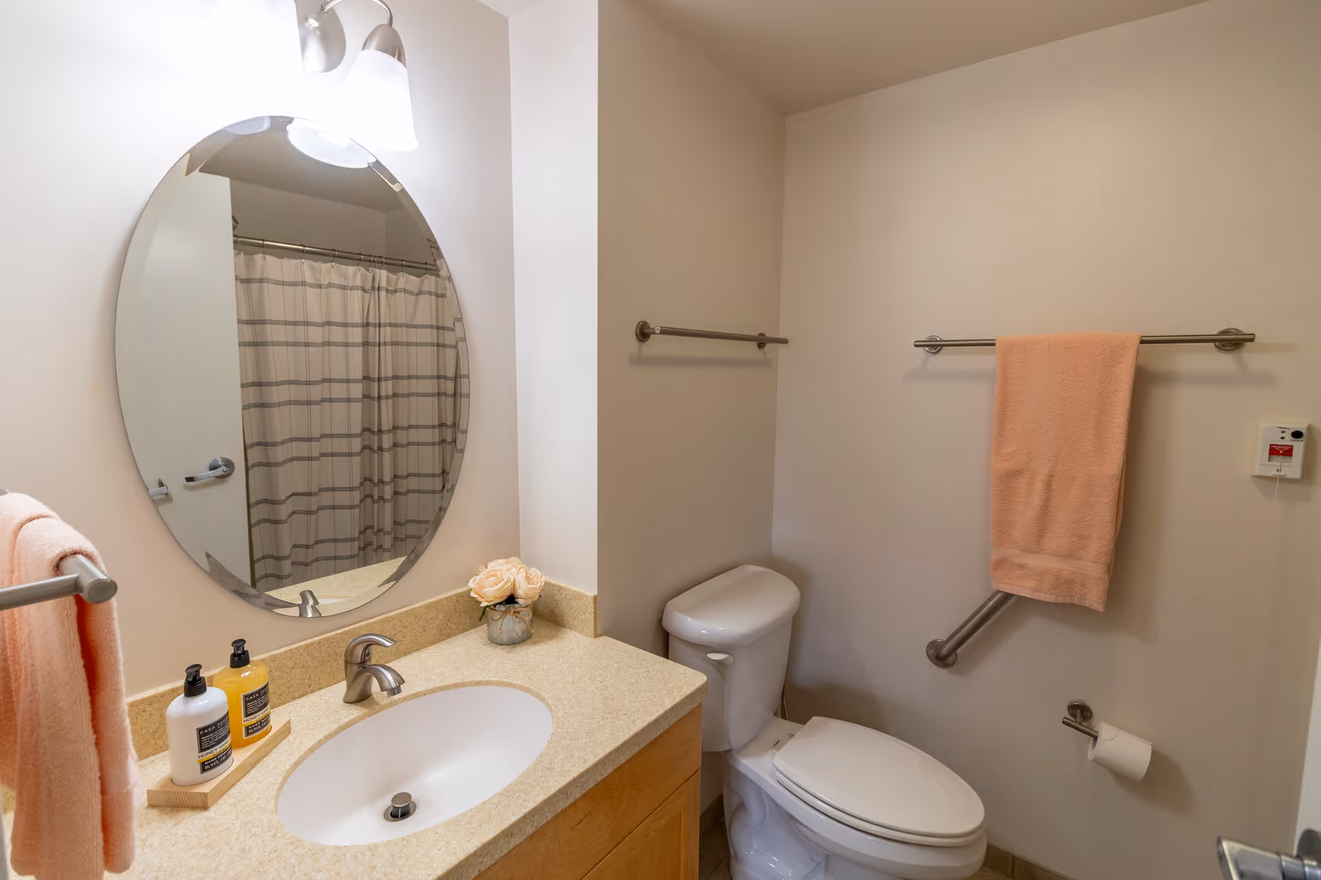 Clean bathroom with a sink and countertop, oval mirror, toilet, shower curtain reflected in the mirror, and peach-colored towels on towel bars.