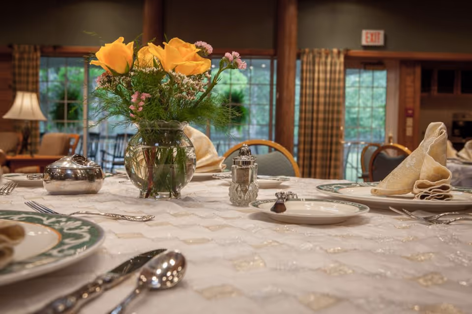 A dining table set with a white patterned tablecloth, plates with folded beige napkins, silverware, a salt shaker, and a small silver dish. A glass vase with yellow roses and small pink flowers is placed in the center. The background shows large windows with checkered curtains and a cozy interior with wooden elements.