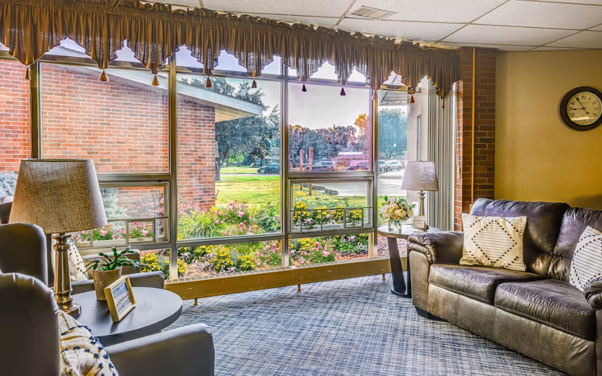 Cozy seating area with a leather sofa, armchairs and lamps in front of large windows overlooking a landscaped garden.