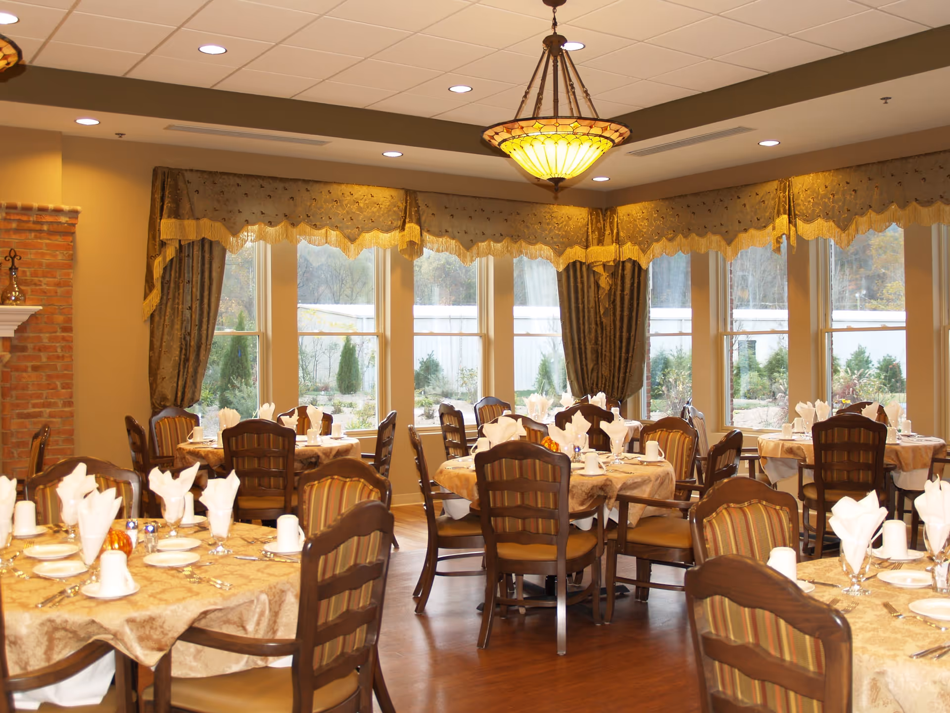 A dining room with multiple round tables covered with beige tablecloths, each set with white napkins, plates, cups, and silverware. The room has large windows with decorative curtains, wooden chairs with striped cushions, a brick fireplace on the left, and a hanging chandelier light fixture.