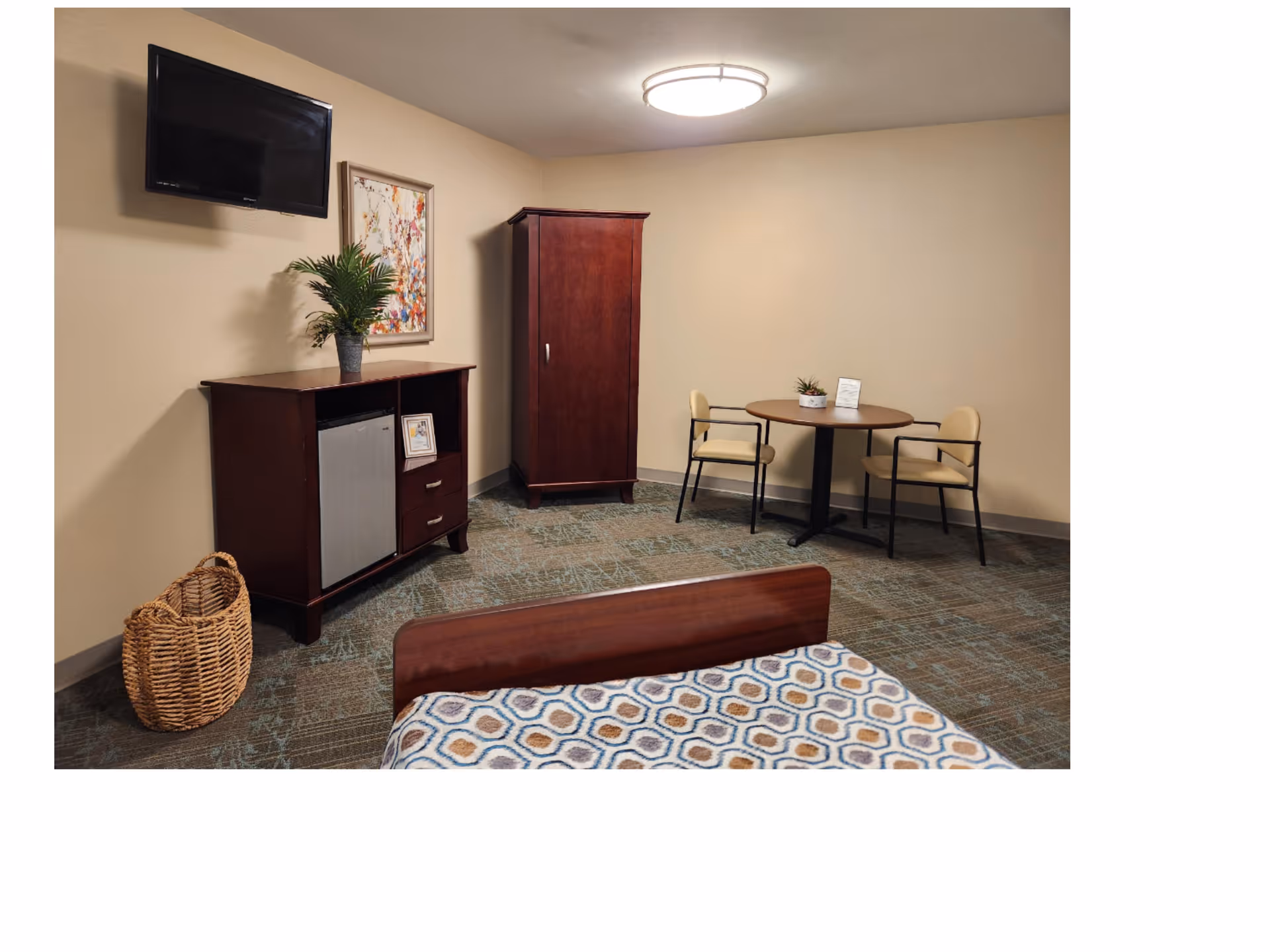 Interior view of a retirement village room featuring a bed with a patterned blanket in the foreground, a wooden cabinet with a small refrigerator and a potted plant on top, a wall-mounted flat-screen TV, a framed artwork, a tall wooden wardrobe, and a round table with two chairs against a beige wall.