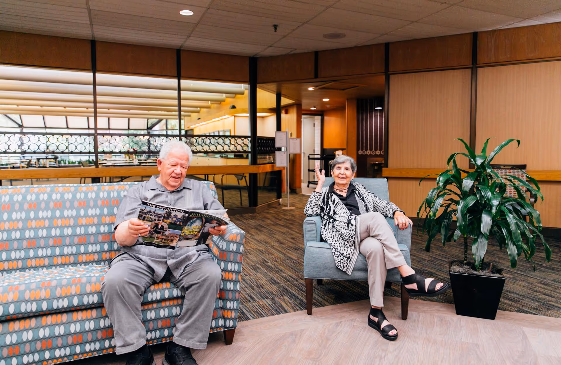 An elderly man and woman sitting in a senior living facility common area. The man is seated on a patterned couch reading a magazine, while the woman sits on a single armchair next to a potted plant. The room has carpeted flooring, wood-paneled walls, and large windows in the background.
