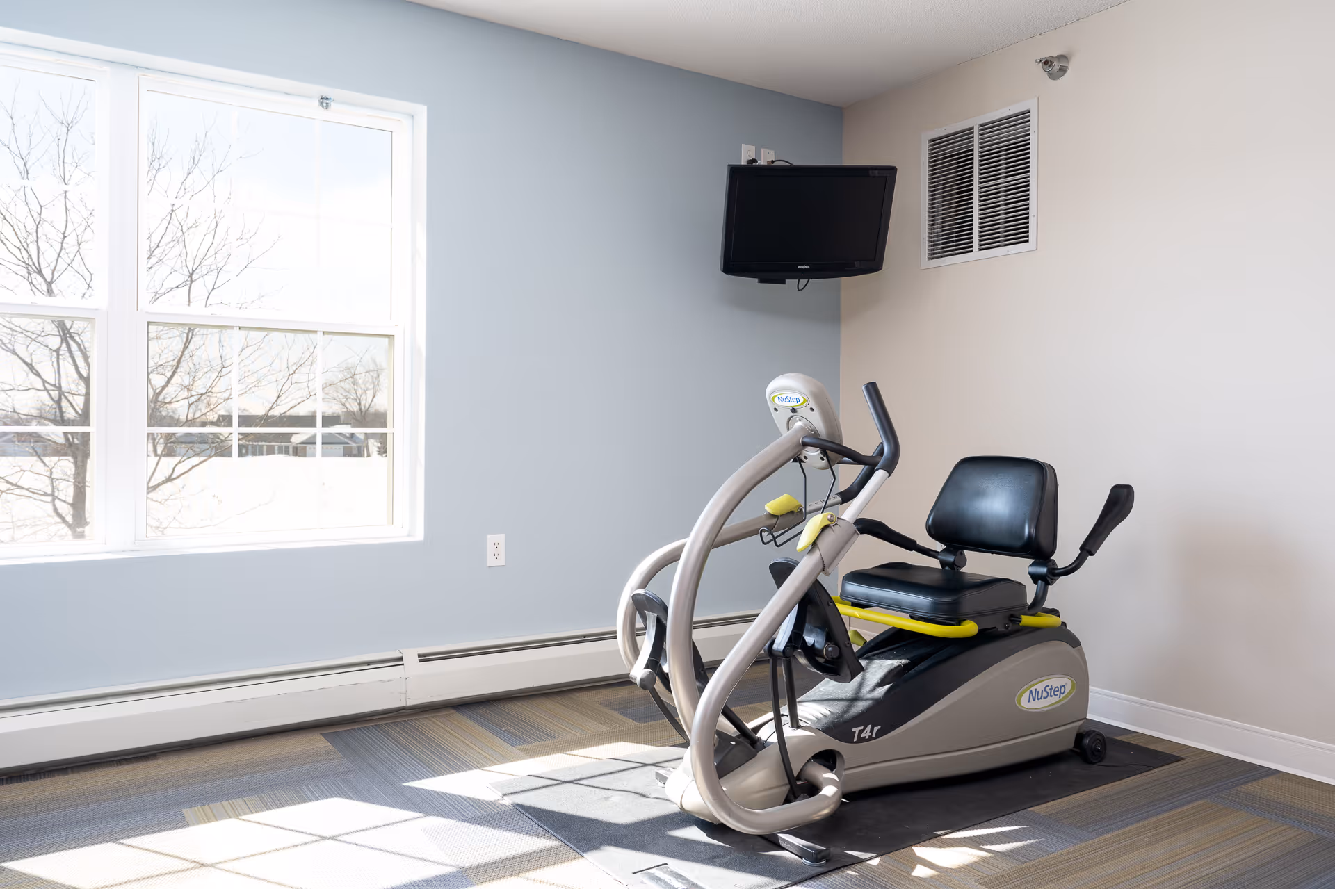 A small exercise room with a NuStep T4r recumbent cross trainer positioned on a mat. The room has light blue and beige walls, a large window letting in natural light, a wall-mounted TV, and a ventilation grille.