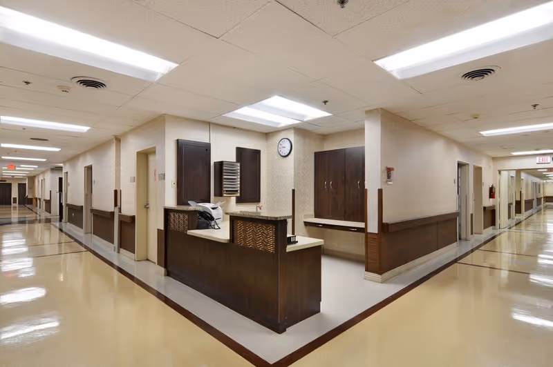 A clean, well-lit hallway in a senior living facility with a central nurse's station featuring a dark wood counter and cabinets. The hallway has beige walls with wood paneling and multiple doors leading to rooms on both sides. A clock is mounted on the wall above the nurse's station, and fluorescent lights illuminate the ceiling.