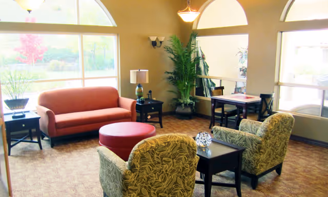 A bright and cozy living room area with a red sofa, two patterned armchairs, a round red ottoman, and several dark wood side tables. The room has large arched windows letting in natural light and a view of greenery outside. There are also potted plants and a small dining table with chairs near the windows.
