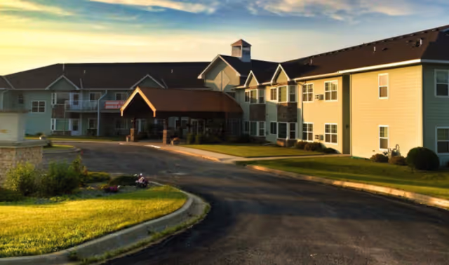Two-story senior living building with a covered entrance, driveway, and landscaped lawn under a warm sky.