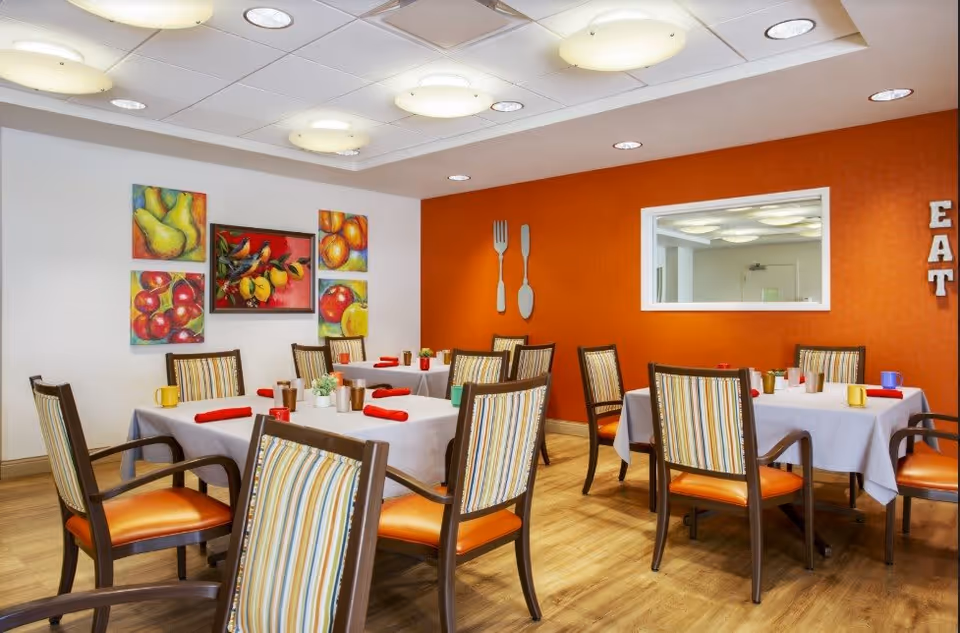 Bright dining room with tables set, striped chairs, an orange accent wall and colorful fruit artwork.