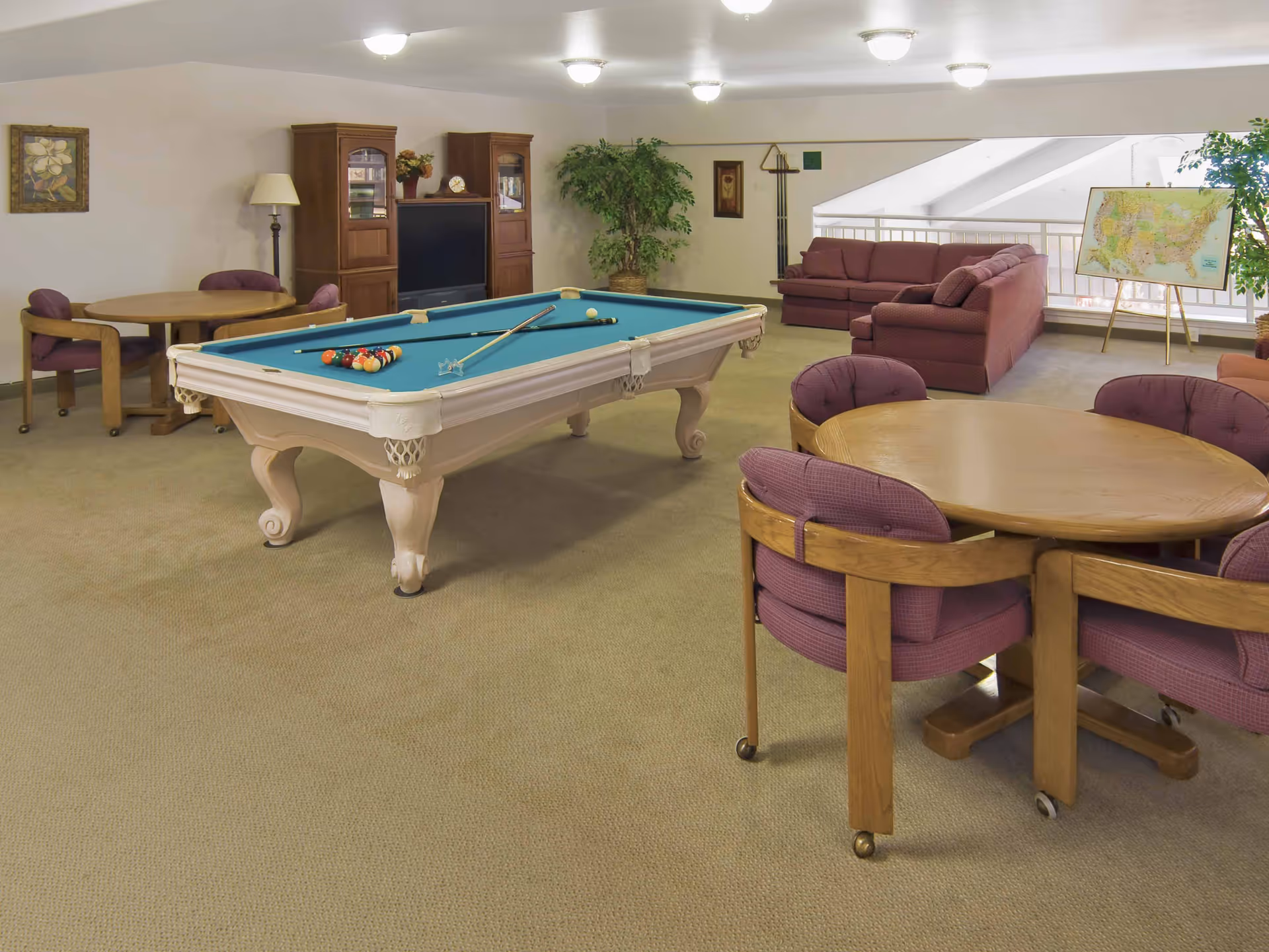 A spacious recreational room with a pool table in the center, two round wooden tables with purple cushioned chairs, a wooden entertainment center with a TV, two maroon sofas, a large potted plant, framed artwork on the walls, and a map on an easel near a railing overlooking a lower level.