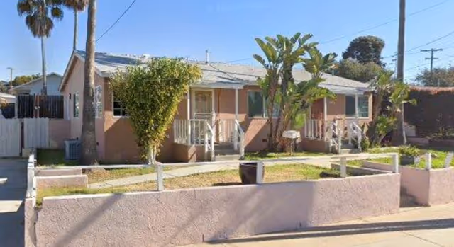 Single-story pink stucco building with small porches, front yard planters and palm trees along a residential street.