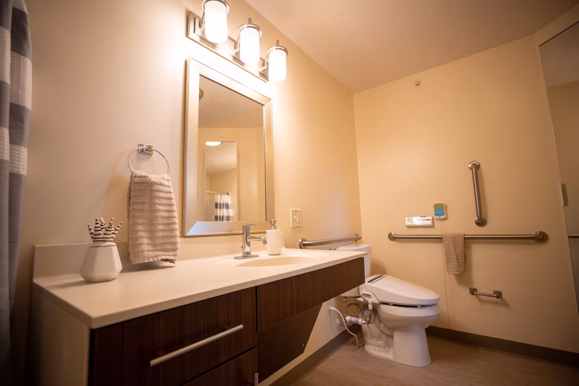 A clean and modern bathroom with beige walls, a white countertop with a sink, a mirror with three lights above it, a towel hanging on a ring, and a toilet equipped with safety grab bars and a bidet attachment.