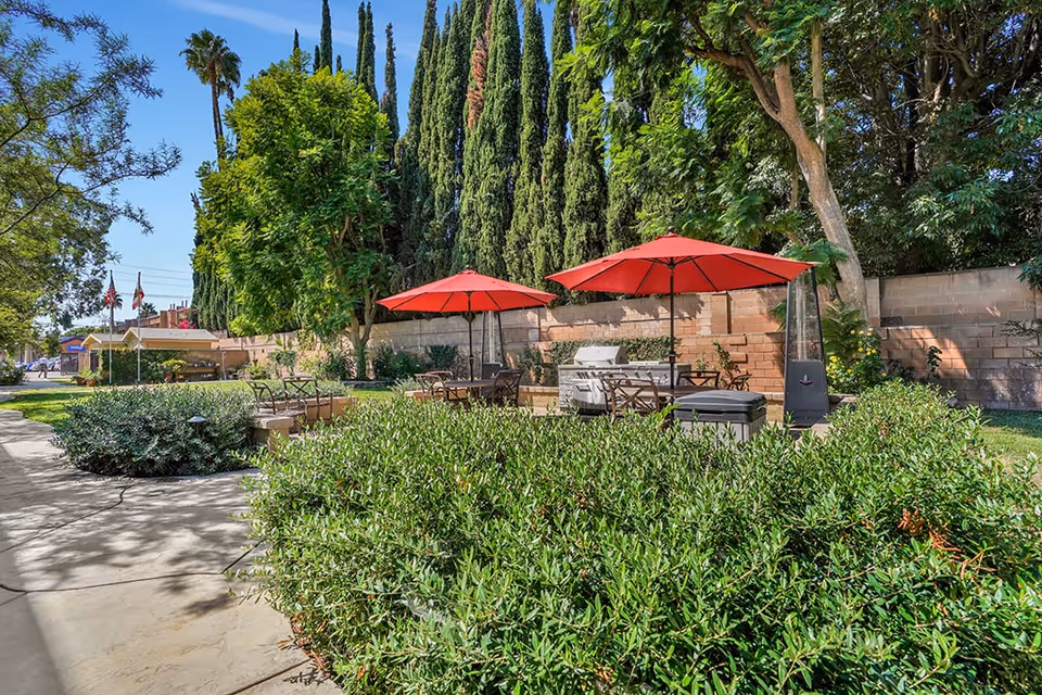 Sunlit outdoor courtyard with red patio umbrellas, tables and chairs next to a barbecue, surrounded by shrubs and tall trees.