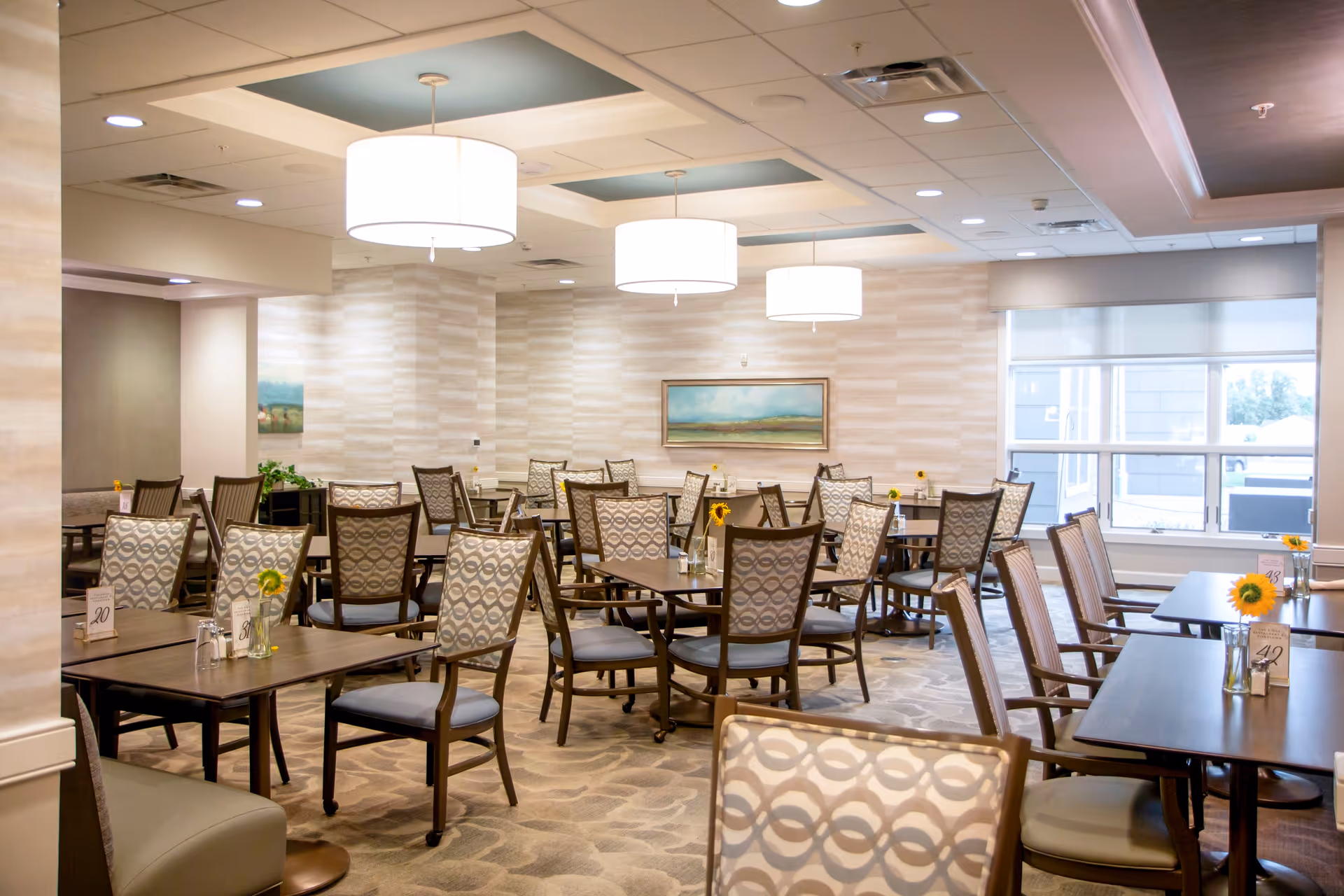 A bright and spacious dining room with multiple wooden tables and patterned cushioned chairs arranged neatly. Each table has a small vase with a sunflower and a table number. The room features large windows allowing natural light, modern ceiling lights, and a landscape painting on the wall.