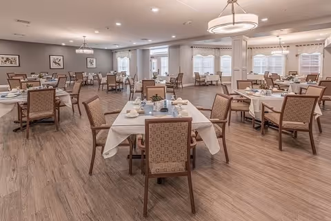A spacious dining room with multiple tables covered with white tablecloths and set with cups, plates, and utensils. The room features wooden chairs with woven backs, large windows with white curtains, and soft lighting from ceiling fixtures. The floor is a light wood finish, and framed artwork is visible on the walls.