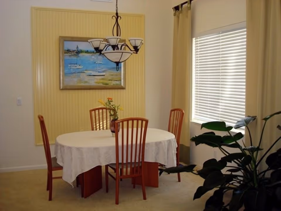 A dining room with a round table covered with a white tablecloth and four wooden chairs around it. A vase with flowers is placed on the table. There is a large window with closed blinds and beige curtains on the right side. A framed painting of boats on water hangs on a yellow paneled wall. A chandelier with multiple lights hangs above the table. A large green plant is visible in the foreground on the right side.
