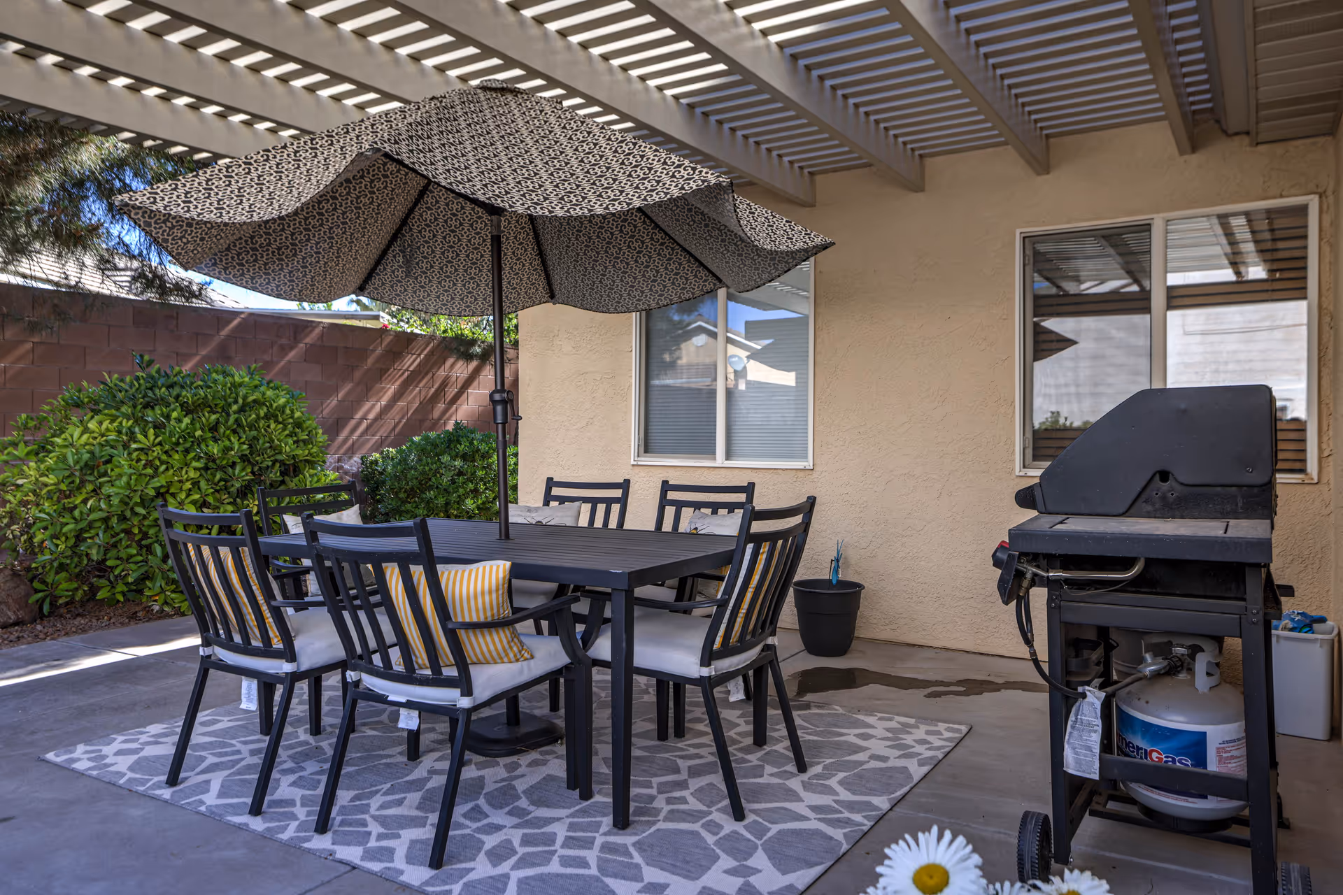 Outdoor patio area with a black dining table and six chairs, some with cushions, under a patterned umbrella. There is a gas grill on the right side and green bushes along a brick wall in the background. The patio is covered with a pergola.