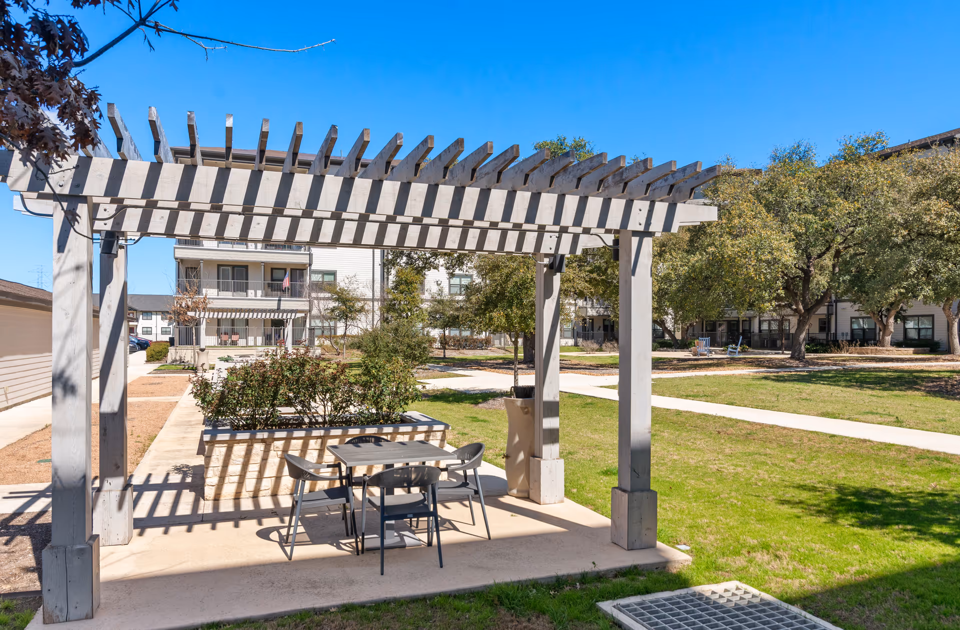 Outdoor courtyard with a wooden pergola shading a table and chairs in front of apartment buildings and trees.