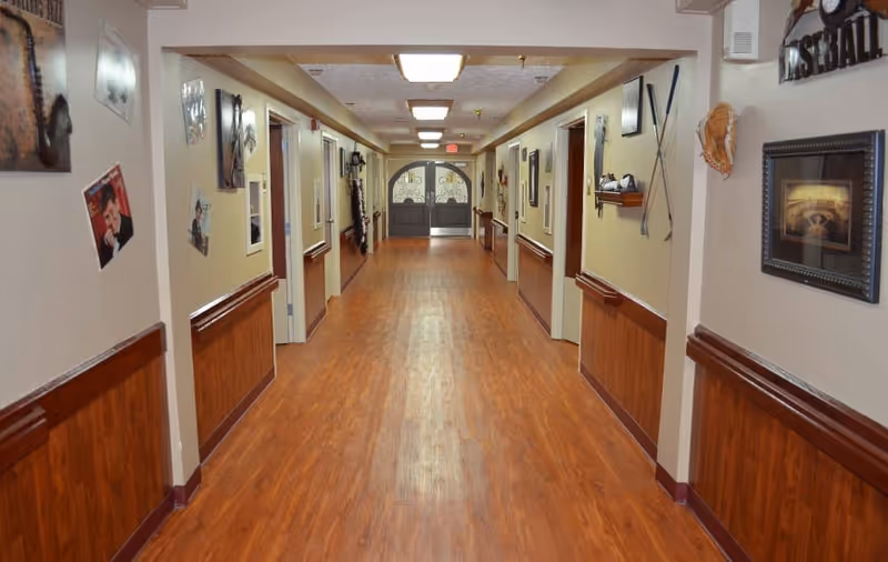 A long hallway in a senior living facility with wood flooring and beige walls. The walls are decorated with various framed pictures and sports memorabilia. There are several doorways along the hallway, and double glass doors are visible at the far end.