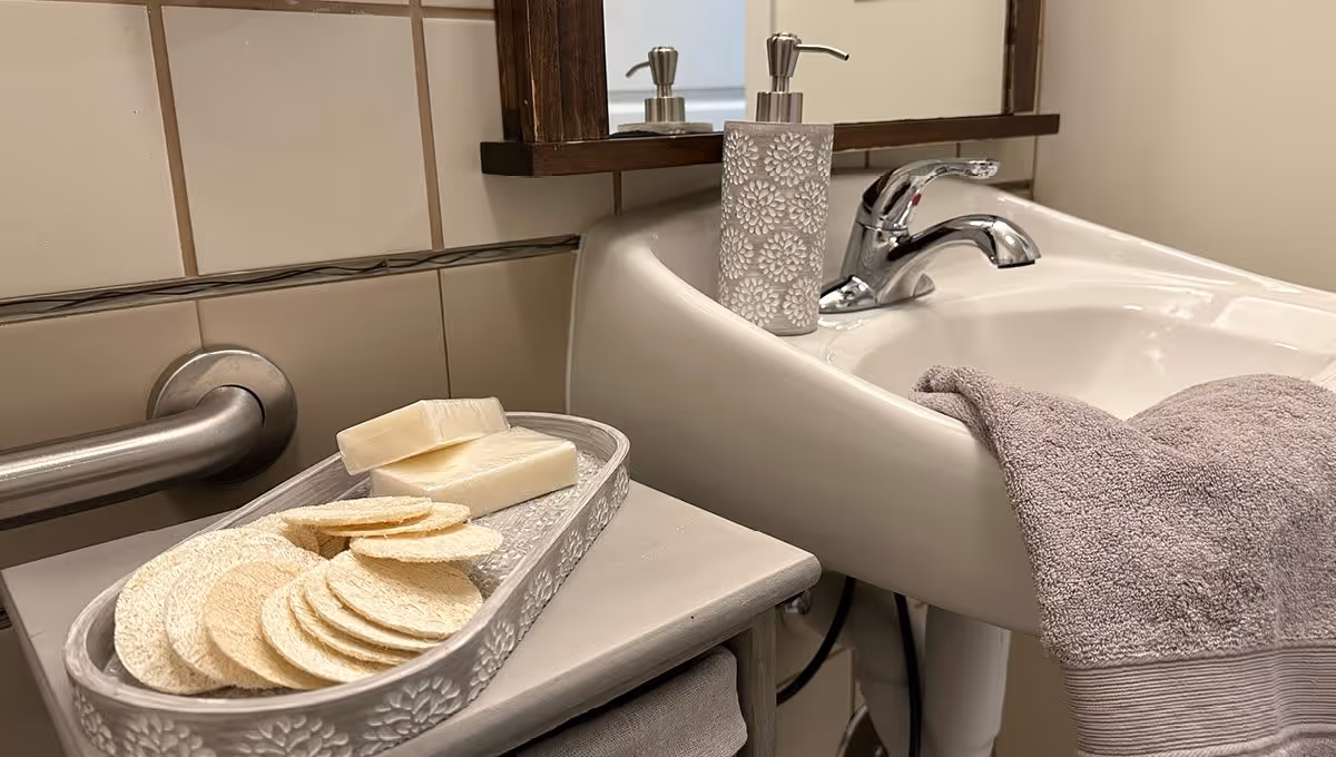 A bathroom sink with a chrome faucet, a lavender towel draped over the edge, a decorative soap dispenser, and a tray holding two bars of soap and several round exfoliating pads. The wall behind has beige tiles and a metal grab bar.