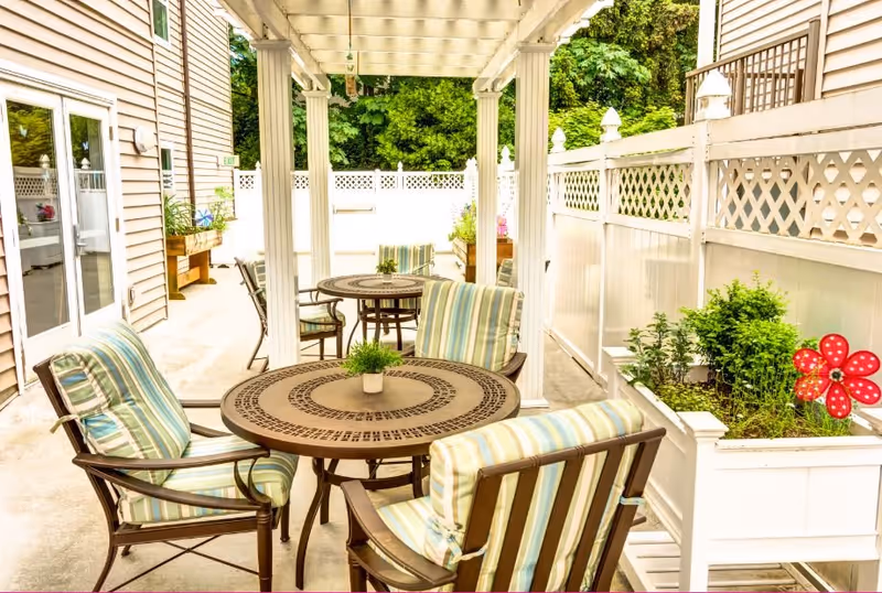 Outdoor patio area at a senior living facility with round metal tables and cushioned chairs featuring striped upholstery. The space is covered by a white pergola and surrounded by white fencing with lattice design. There are planter boxes with greenery and a decorative red pinwheel flower.