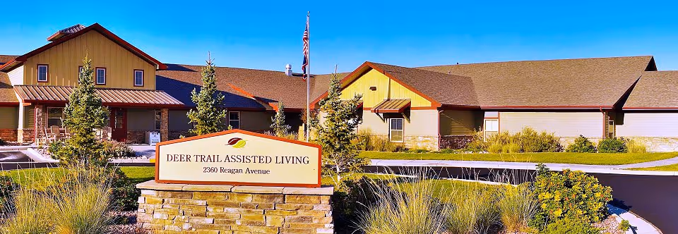 Exterior view of Deer Trail Assisted Living facility with a stone sign in front displaying the name and address 2360 Reagan Avenue. The building has a brown roof, beige and yellow walls, and is surrounded by landscaped greenery and trees under a clear blue sky.