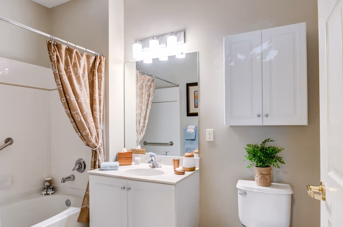 A clean and well-lit bathroom featuring a white bathtub with a beige shower curtain decorated with a white leaf pattern. There is a white vanity with a sink, a large mirror above it, and a light fixture with four bulbs. On the vanity are a wooden tissue box, a small towel, and decorative items. A white cabinet is mounted on the wall above a white toilet, which has a green potted plant on top. The walls are painted a light beige color.