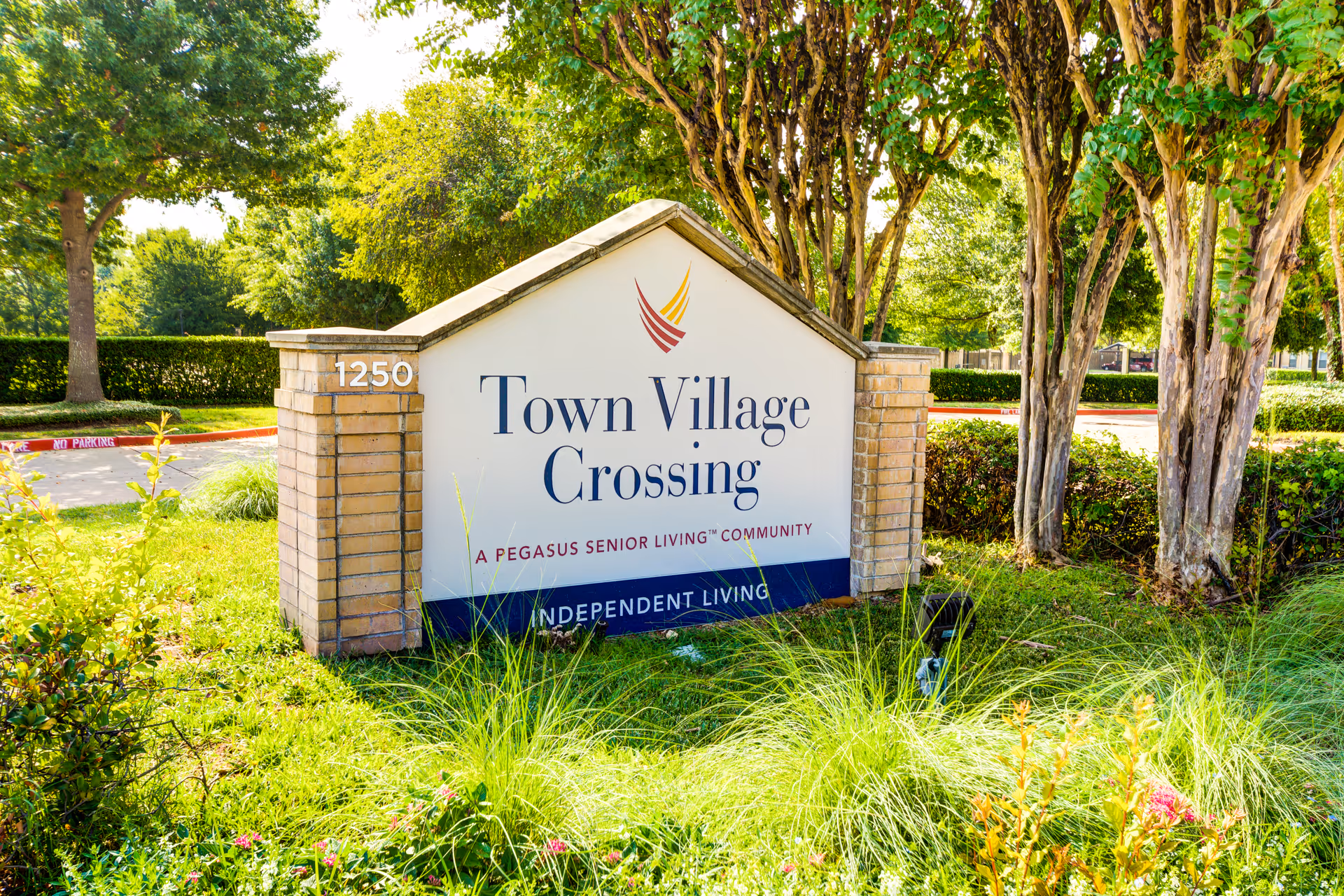 Stone entrance sign reading "Town Village Crossing" and other community details amid trees and landscaped grass.
