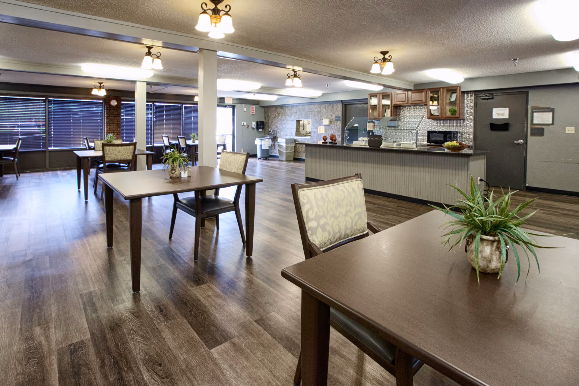 A spacious dining area with several wooden tables and chairs, each table decorated with a small potted plant. The room has large windows with blinds, wood flooring, and ceiling lights. In the background, there is a kitchen area with cabinets, a microwave, and a countertop.
