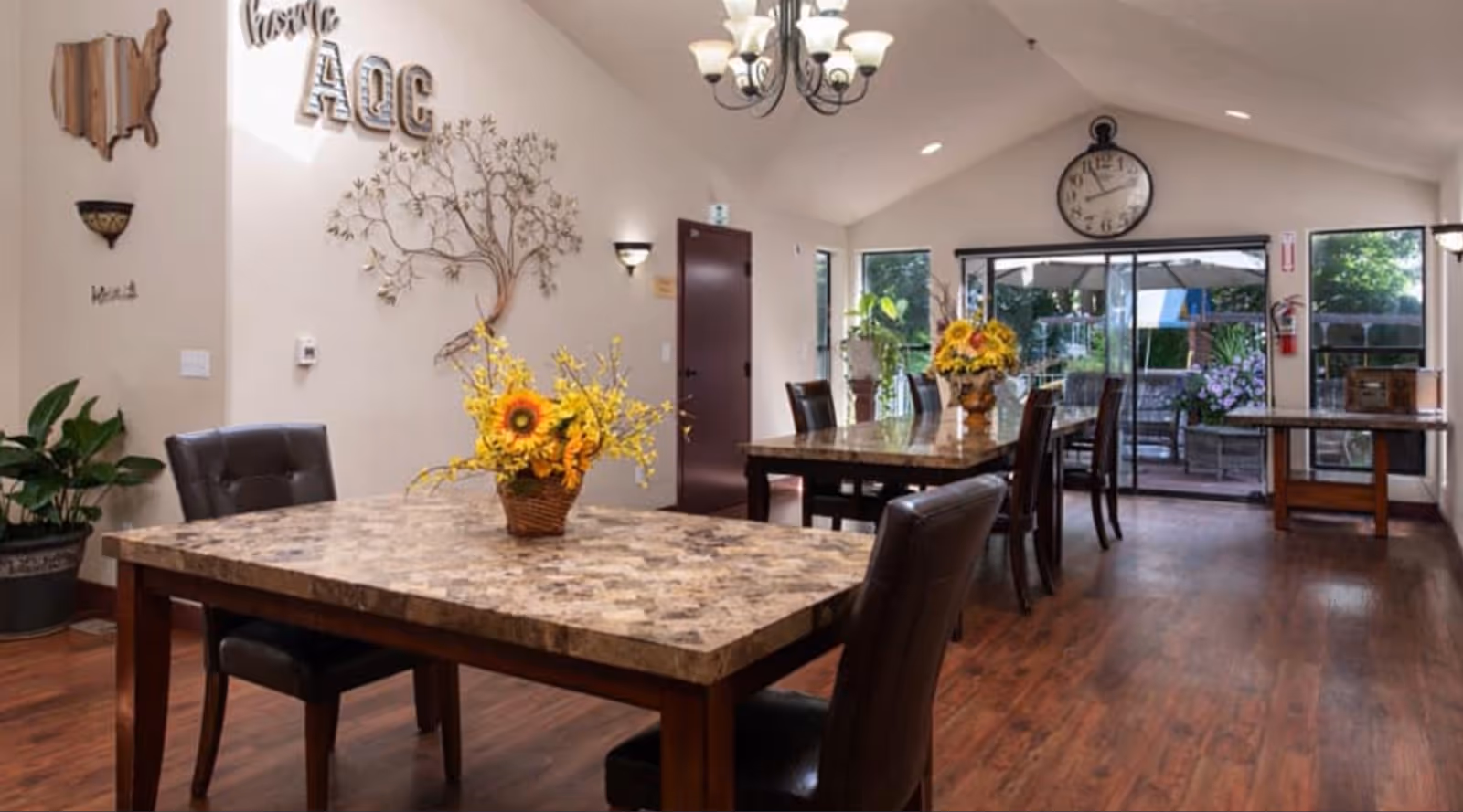Interior view of a dining room with two marble-top tables, each decorated with a vase of yellow flowers. The room has wooden flooring, leather chairs, a large wall clock, and sliding glass doors leading to an outdoor patio area. Wall decorations include a metal tree sculpture, a wooden map of the United States, and the letters 'AQC'.