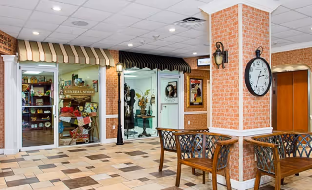 Interior view of a senior living facility hallway with a tiled floor, brick-patterned walls, and a large clock mounted on a pillar. There are two wooden benches with decorative backs placed near the pillar. On the left side, there are storefront-style entrances labeled 'General Store' and another shop with awnings above the doors. The ceiling has recessed lighting and a white grid pattern.