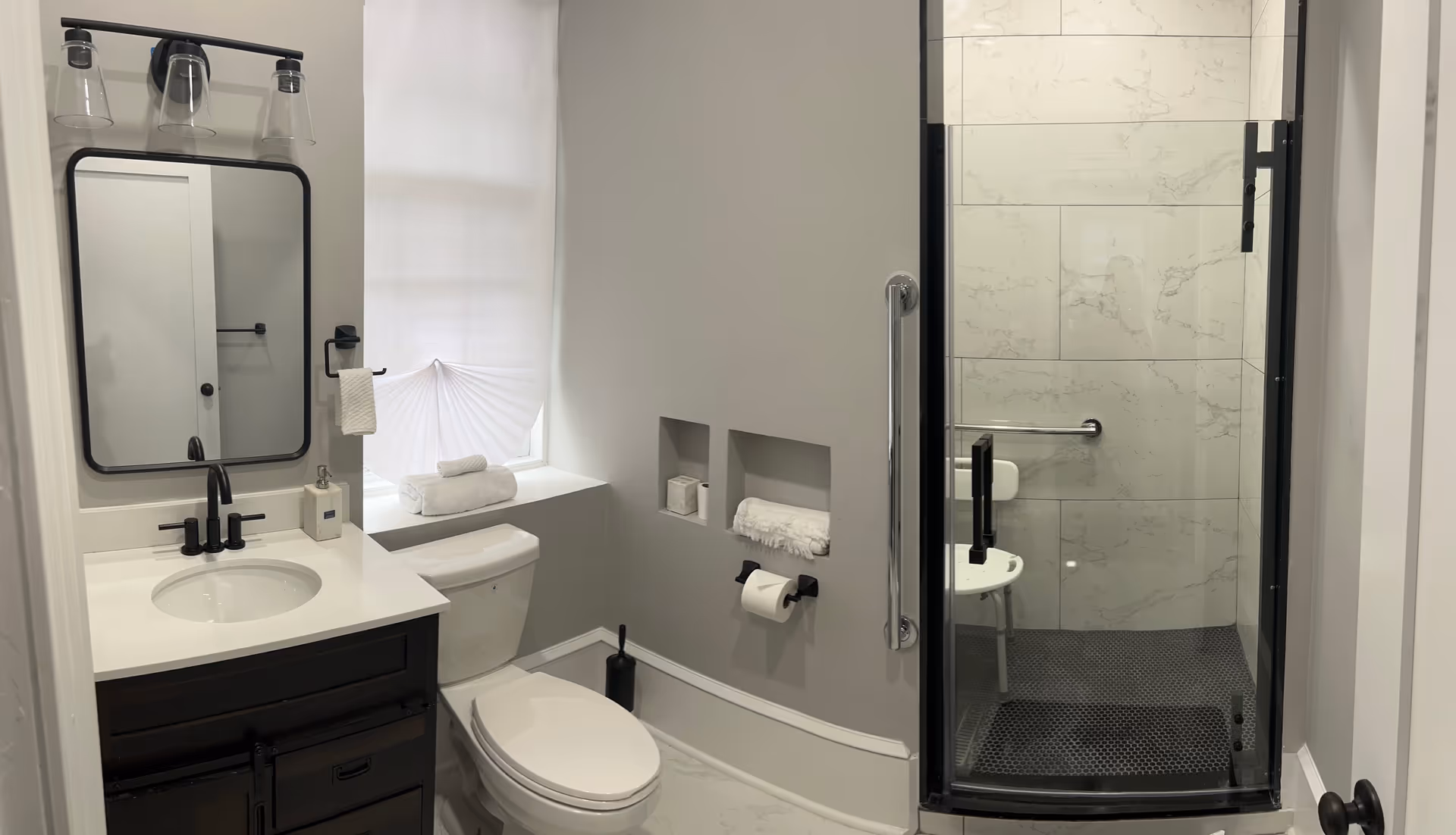 A modern bathroom featuring a white toilet, a dark wood vanity with a white countertop and sink, a rectangular mirror with black frame, and a three-light fixture above the mirror. There is a window with a white pleated shade, built-in wall niches holding towels and toilet paper, and a glass-enclosed shower with marble-patterned tiles and a white shower chair inside.