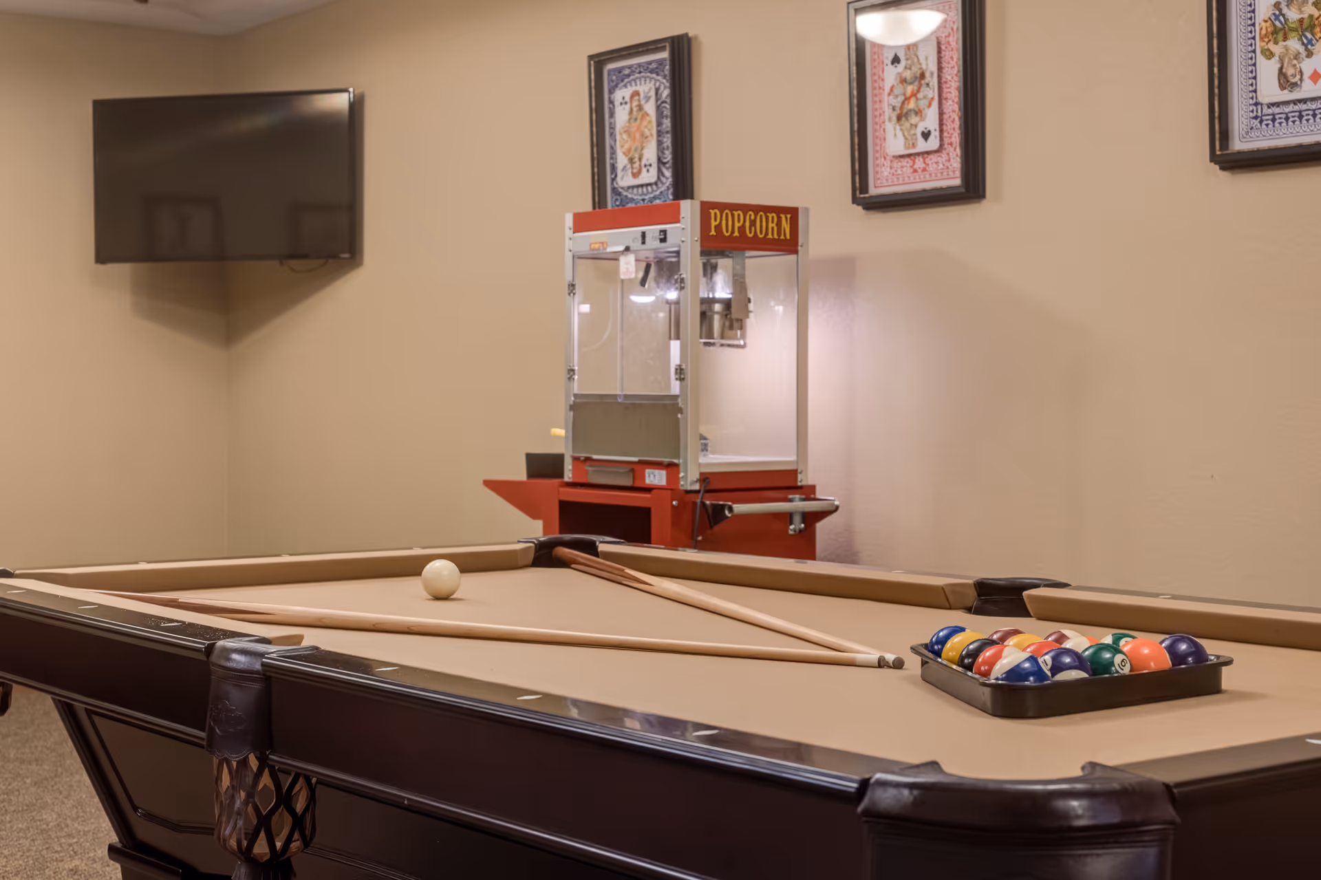 A game room with a pool table featuring a beige felt surface, two pool cues, a white cue ball, and a rack of colorful billiard balls. In the background, there is a red popcorn machine and a wall-mounted flat screen TV. The walls are decorated with framed playing card artwork.