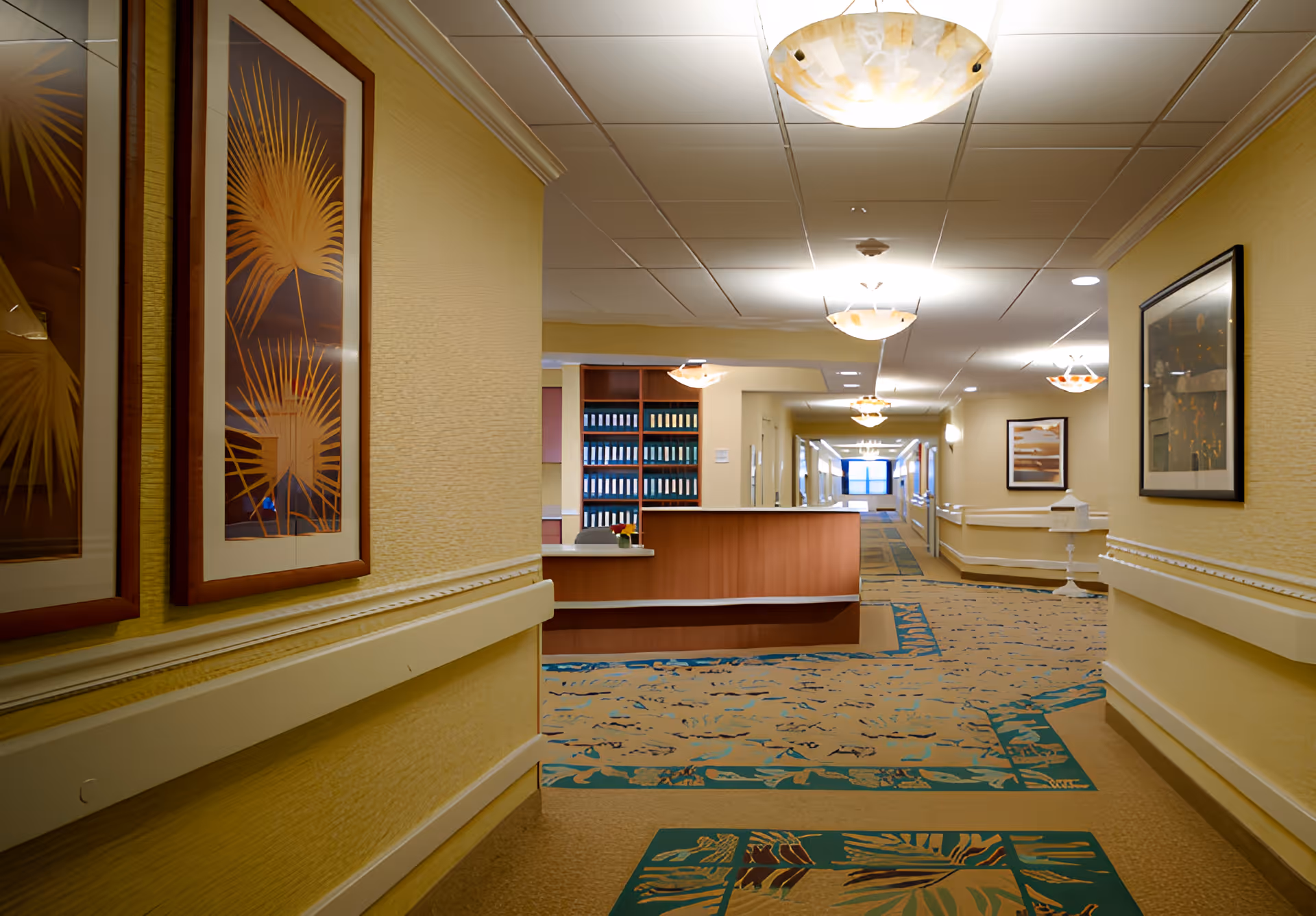 A well-lit hallway in a senior living facility with yellow walls and patterned carpet. The hallway features a wooden reception desk with shelves of binders behind it. Framed artwork hangs on the walls, and ceiling lights provide warm illumination.
