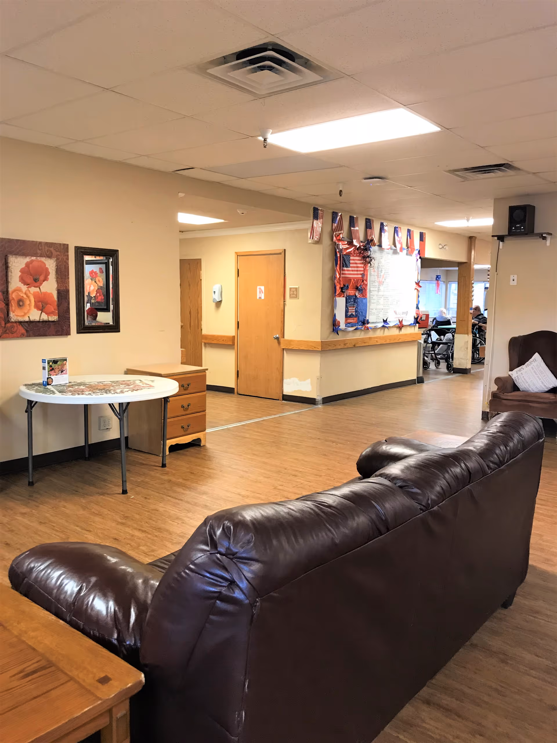 Interior view of a nursing home common area with a brown leather couch in the foreground, a round table with a floral tablecloth and a small wooden drawer unit against the wall. The walls are beige with two framed floral paintings. In the background, there is a hallway with wooden doors and a bulletin board decorated with American flags and red, white, and blue decorations. Some people in wheelchairs are visible further down the hallway.