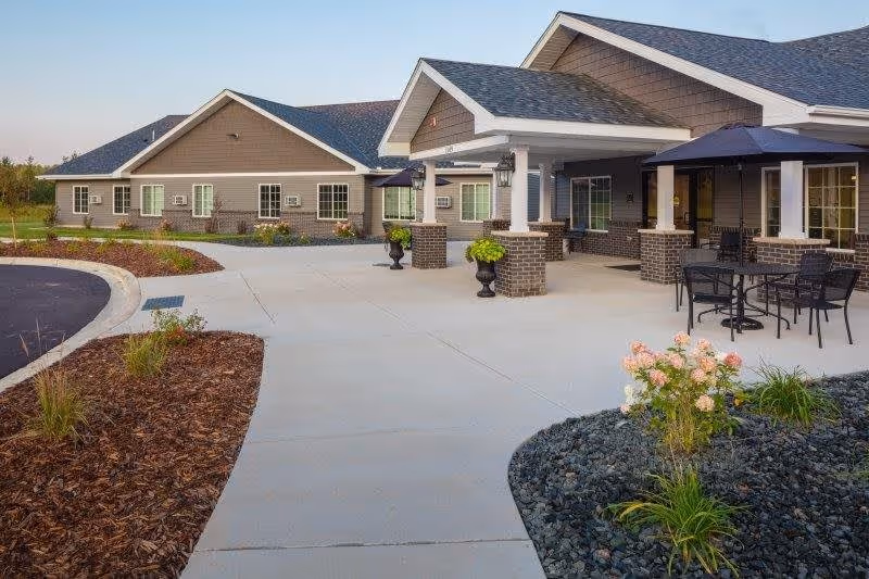 Exterior view of a senior living facility with a wide concrete walkway, landscaped garden beds with flowers and shrubs, and a covered patio area with tables and chairs. The building has brown siding, white trim, and multiple windows under a clear sky.