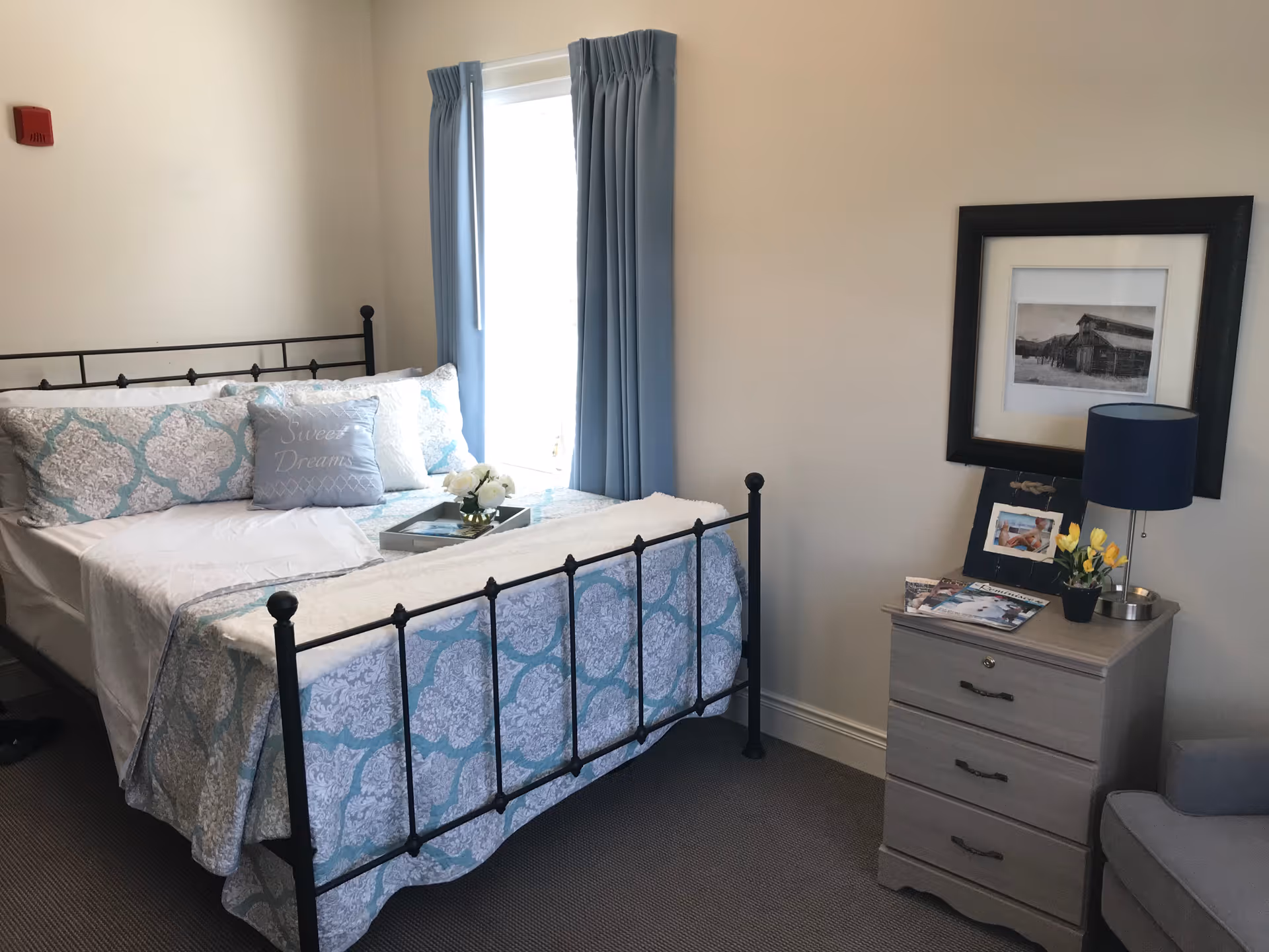 A cozy bedroom with a black metal bed frame, a bed made with white and light blue patterned bedding, and a pillow that says 'Sweet Dreams'. There is a window with light blue curtains letting in natural light. Next to the bed is a light gray nightstand with three drawers, a blue lampshade, a small vase with yellow flowers, a framed photo, and some magazines. A framed black and white picture hangs on the wall above the nightstand.