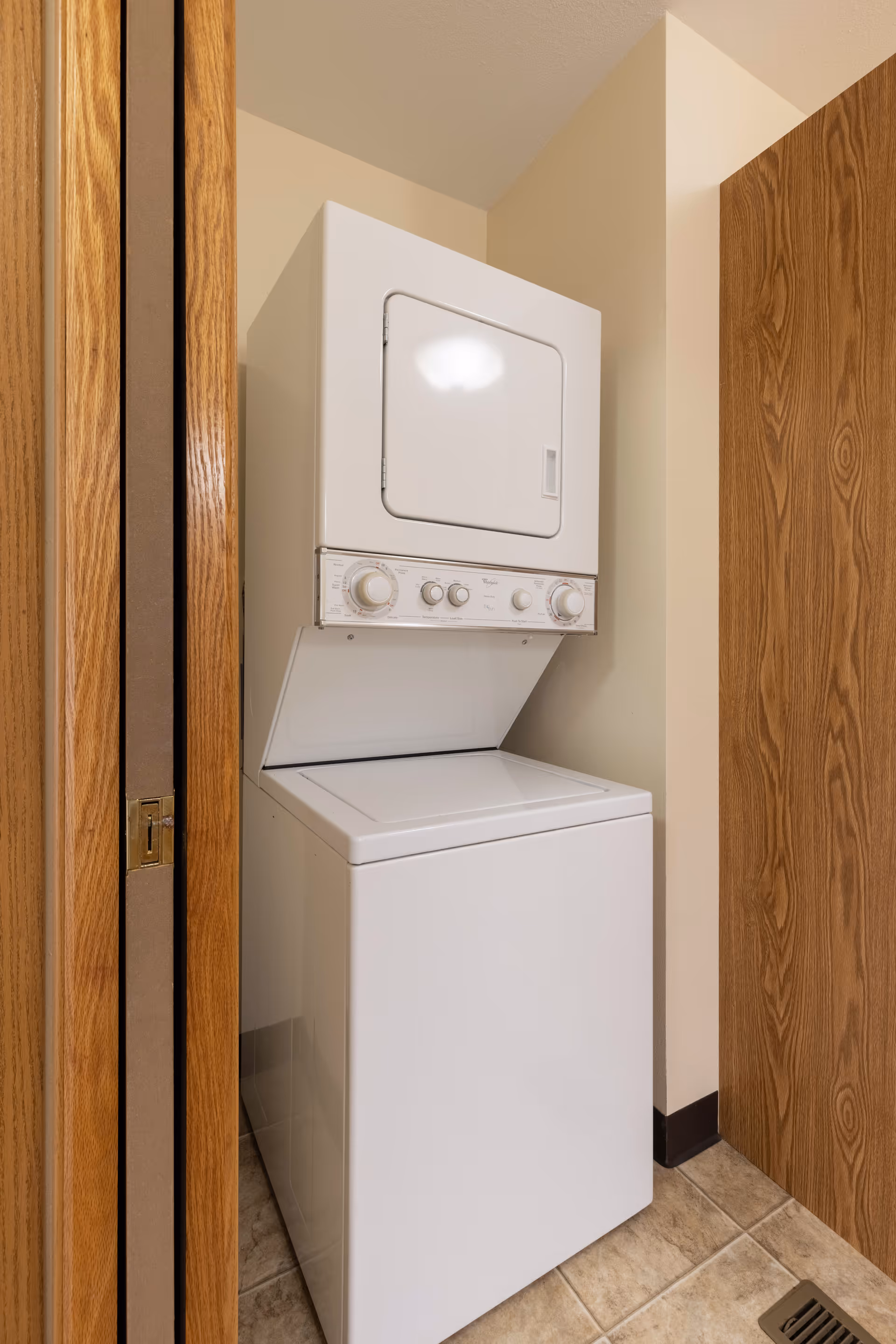 A compact white stacked washer and dryer unit placed in a small laundry nook with beige walls and tiled floor, flanked by wooden doors.