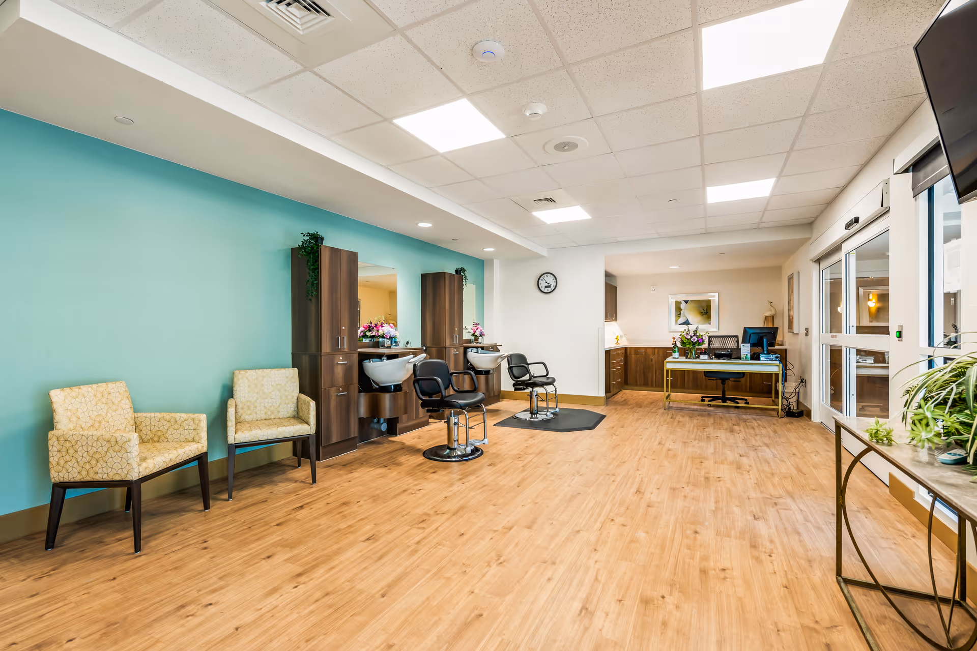Interior view of a salon area in a senior living facility with two salon chairs and wash basins against a teal wall, two patterned armchairs, a reception desk with a computer, and large windows letting in natural light.