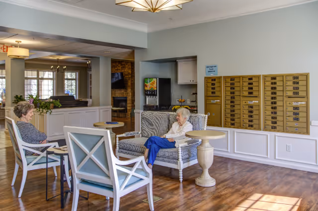 Two elderly women sitting and conversing in a bright, spacious common area with wooden flooring, white and blue cushioned chairs, a small round table, a wall with mailboxes, and a beverage dispenser in the background.