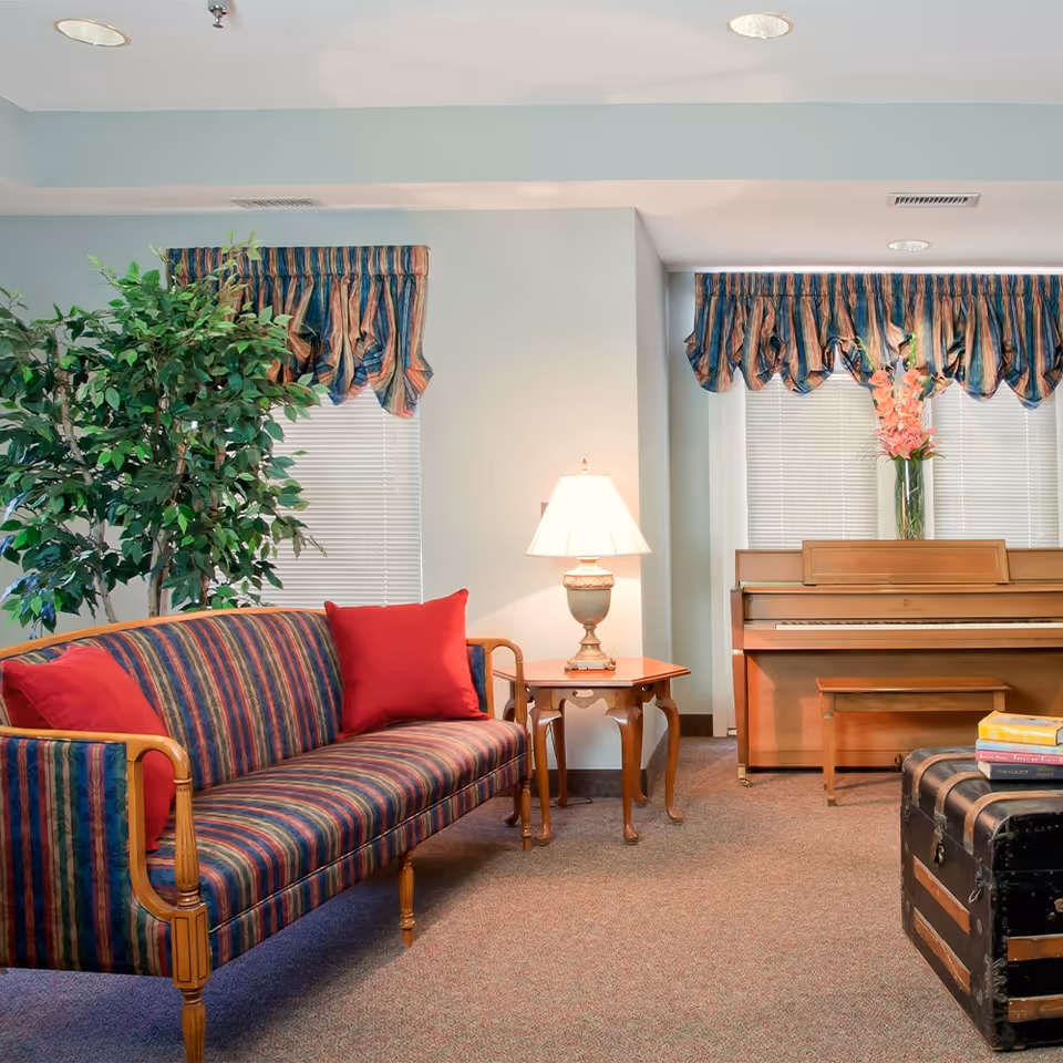 A cozy common room with a striped sofa and red pillows, a side table with a lamp, an upright piano, and a potted plant.