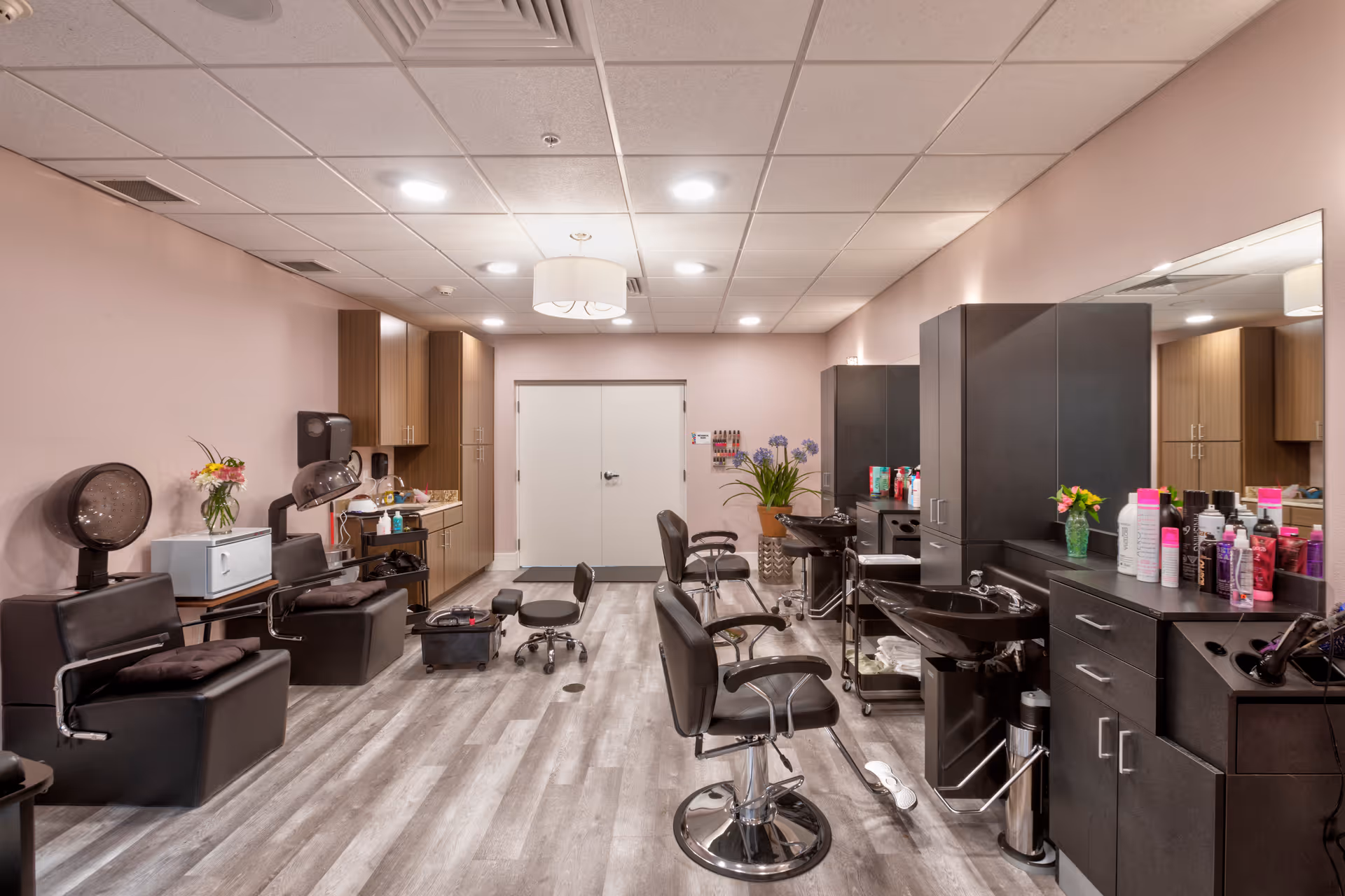 Interior view of a hair salon with multiple black styling chairs, hair washing sinks, and hair care products on counters. The room has light pink walls, wood cabinets, and a light gray wood floor. There are flowers in vases placed on counters and a double door at the far end of the room.
