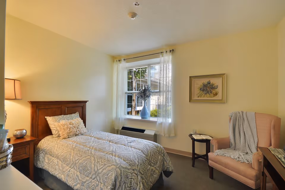 A cozy bedroom with a single bed covered in a patterned comforter and decorative pillows. Next to the bed is a wooden nightstand with a lamp and a small decorative item. A window with sheer white curtains lets in natural light, and a vase with flowers sits on the windowsill. Across from the bed is a pink upholstered armchair draped with a gray blanket, beside a small round wooden table holding a glass and a book. A framed floral artwork hangs on the wall above the chair.