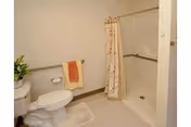 A clean and simple bathroom featuring a white toilet with a plant on the tank, a towel rack with an orange and yellow towel, a white bath mat, and a walk-in shower with a floral shower curtain.