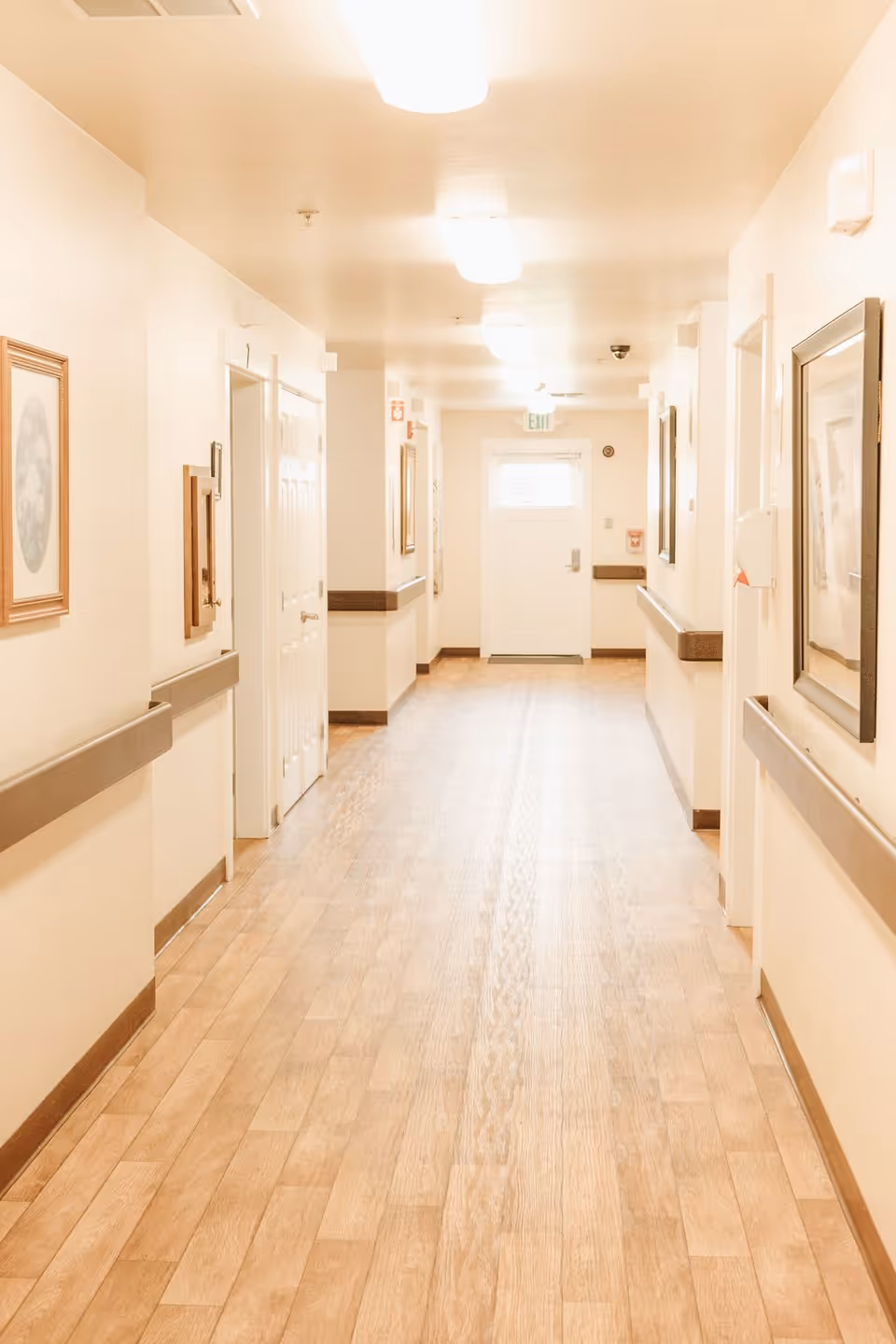 A well-lit indoor hallway with light wood flooring and cream-colored walls. The hallway has handrails on both sides and framed pictures hanging on the walls. Several doors line the hallway, and there is an exit door at the far end with an illuminated exit sign above it.