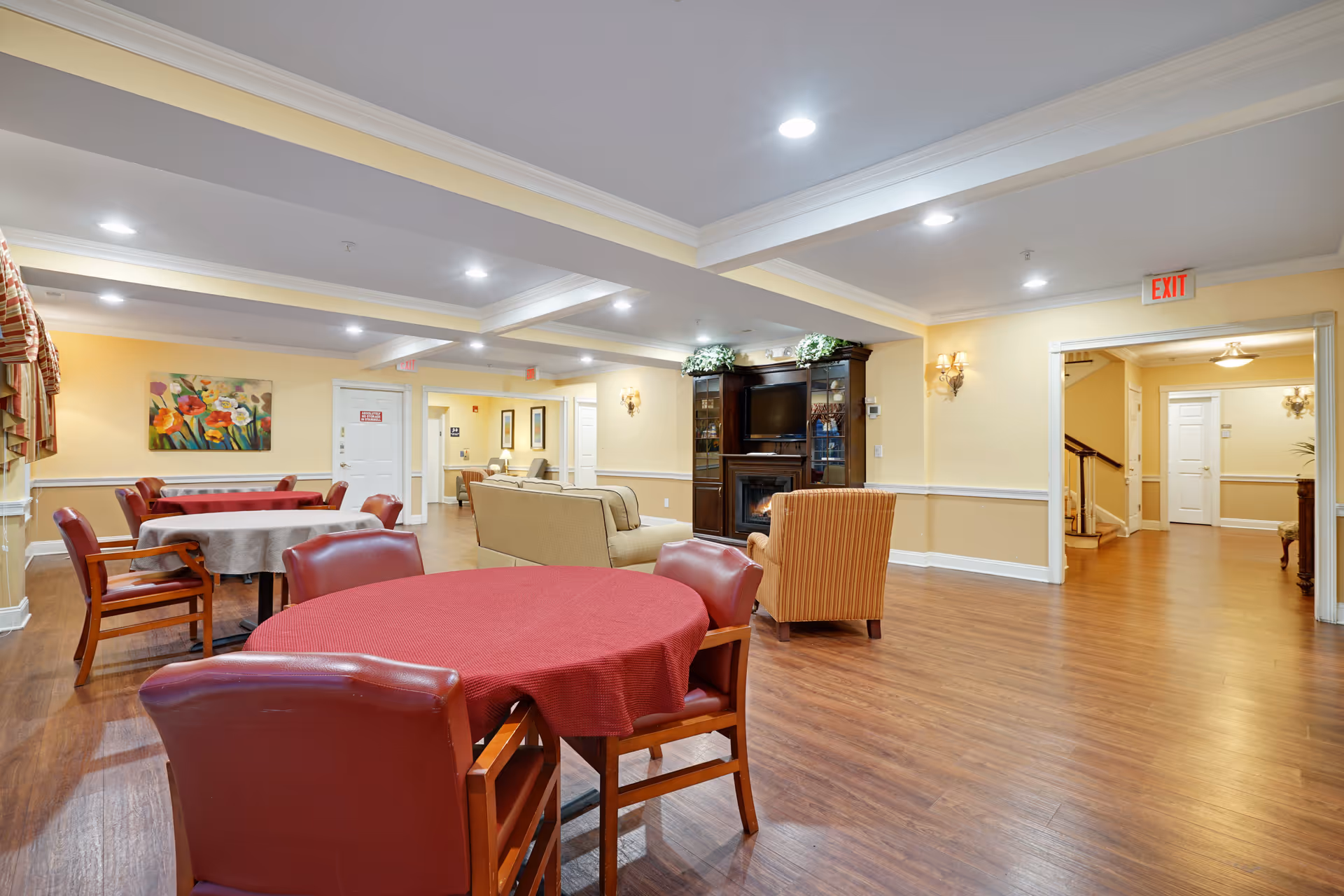 A spacious common area in a senior living facility with several round tables covered with tablecloths and surrounded by chairs. There is a seating area with a beige sofa and an orange armchair facing a dark wood entertainment center with a TV and decorative plants. The room has wood flooring, soft yellow walls, recessed lighting, and an exit sign above a doorway.