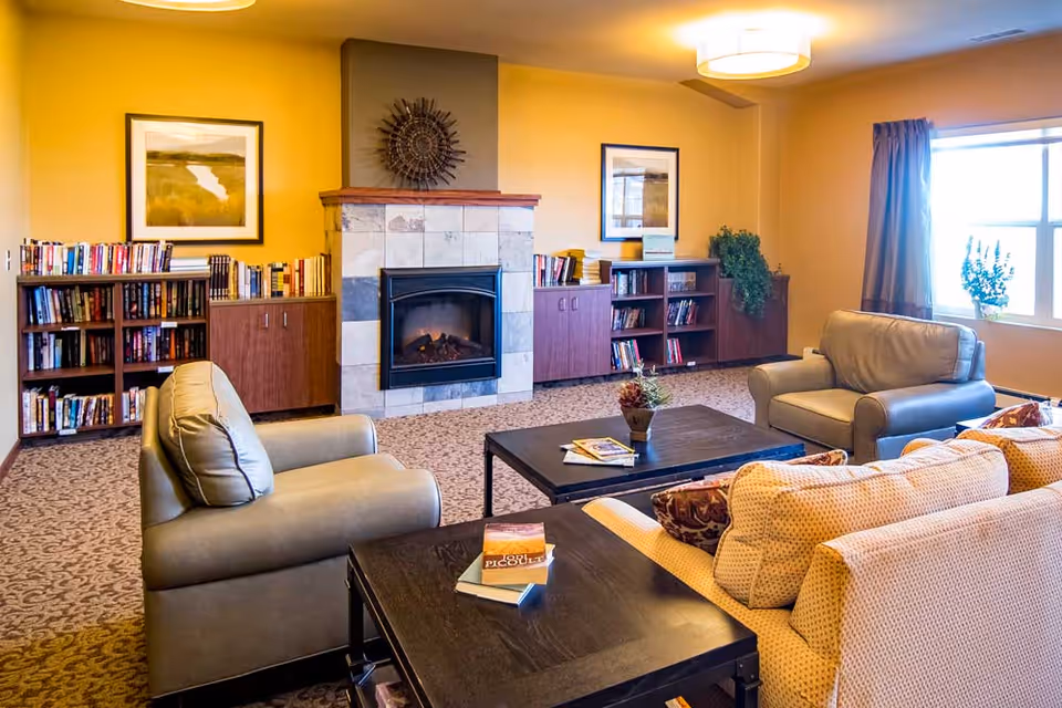 A cozy living room area with beige walls and carpeted floor featuring a fireplace with a decorative piece above it. There are bookshelves filled with books on either side of the fireplace. The room has comfortable seating including a beige sofa and two armchairs arranged around a black coffee table with books and a small plant. A window with curtains lets in natural light.