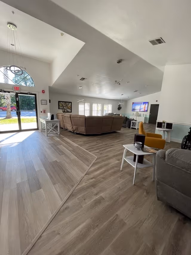 A spacious and well-lit living room area in a senior living facility with large windows covered by white shutters. The room features a large brown sectional sofa, a yellow armchair, a gray recliner, and several small white side tables. A flat-screen TV is mounted on the far wall, and modern pendant lights hang from the ceiling. The floor is covered with light wood-style vinyl flooring, and there is a glass door entrance with an exit sign above it.