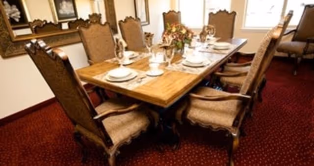 A formal dining room with a wooden table set for six people. The table is adorned with white plates, cups, and glassware, along with a floral centerpiece. The chairs around the table are upholstered with a classic design. The room has a red carpet and framed artwork on the walls.