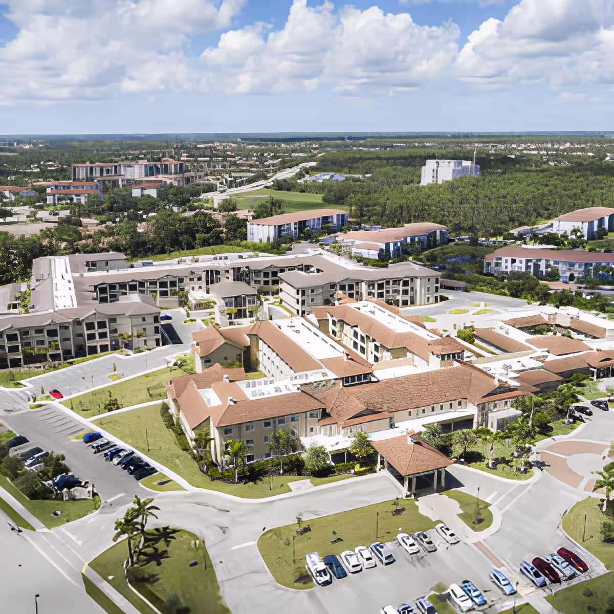Aerial view of the Discovery Village at Naples campus showing multiple connected buildings with red-tiled roofs, parking lots, driveways, and surrounding greenery.