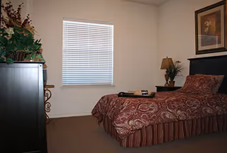 A bedroom with a single bed covered in a red patterned bedspread and a striped bed skirt. There is a dark wooden headboard and a nightstand with a lamp and a small plant. A framed picture hangs on the wall above the bed. A window with closed blinds is centered on the opposite wall. A dark wooden dresser with a decorative plant on top is visible on the left side of the room.